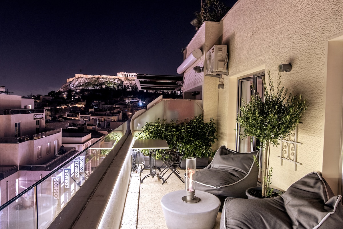 The private roof deck features modern seating arrangements, with a small table and comfortable chairs. Soft lighting enhances the space, while the Acropolis stands majestically in the background, creating a serene atmosphere perfect for relaxation.
