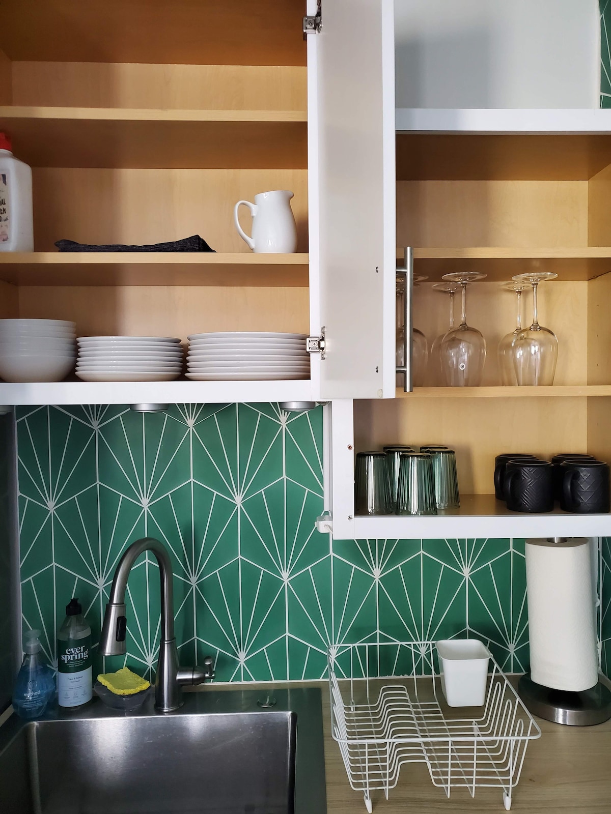Kitchen cabinetry displays a range of dishware, including white plates and glassware, against a patterned green backsplash. A sink with a sleek faucet is positioned beneath the cabinets. Additional items such as a paper towel holder and drying rack are also visible in the space.