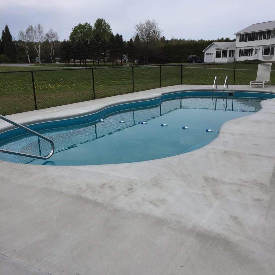 A rectangular pool is set in a fenced area, surrounded by a concrete deck. Several lounge chairs are positioned nearby, and the pool is filled with clear water. Green grass and trees are visible in the background, contributing to a serene outdoor setting.