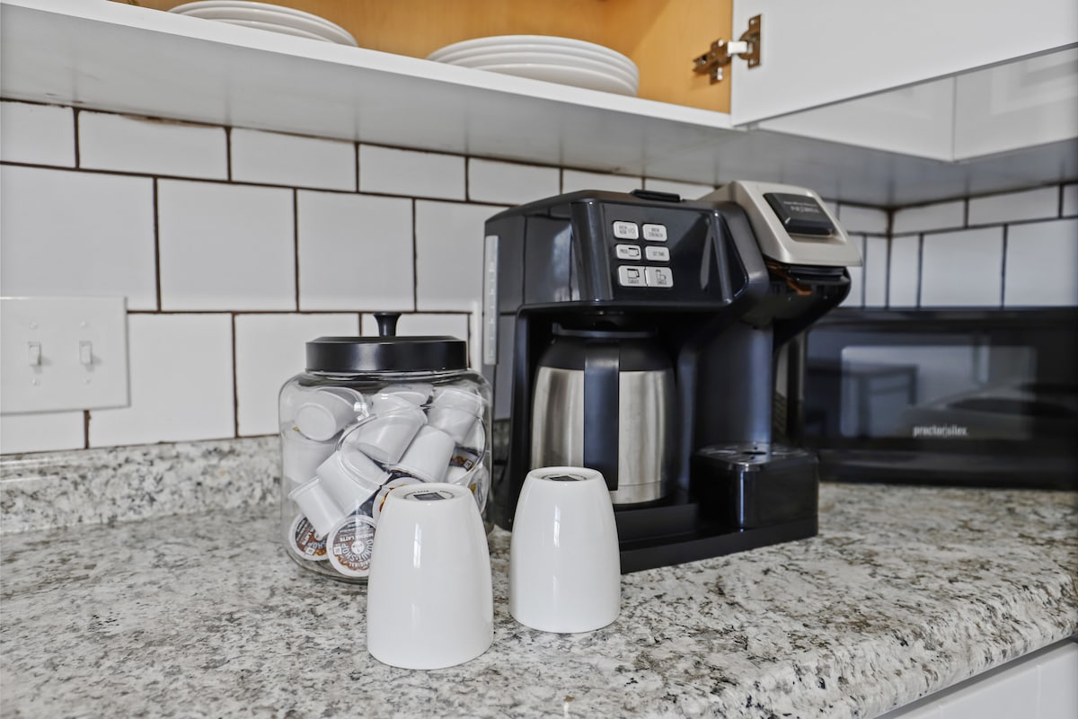 A modern coffee station is set on a speckled granite countertop, featuring a coffee maker with a stainless steel carafe. Next to it, a clear jar is filled with coffee pods, and two white ceramic cups are placed upside down for storage. Shelves above display stacked plates.