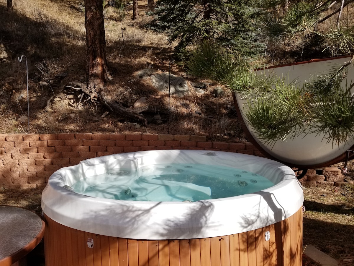 A round hot tub is set on a wooden deck, surrounded by a natural landscape of pine trees and hillside. The tub features a light blue interior, with bubbling water indicating warmth. A small round table can be seen nearby, adding to the outdoor relaxation space.