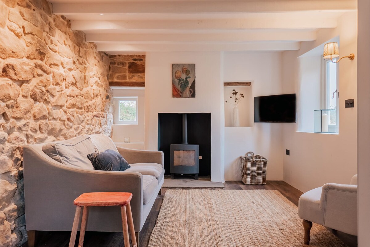 A cozy living area is presented with a stone wall and white plaster accents. A comfortable gray sofa faces a multi-fuel burner, with a small wooden table in front. Light fixtures and large windows allow natural light to enhance the space.