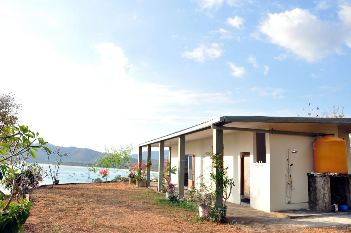 The villa is positioned on a gently sloping landscape, featuring a spacious exterior with a covered porch. Potted plants add greenery, while ocean views extend beyond the building. A water tank is visible on the side, contributing to the natural surroundings.