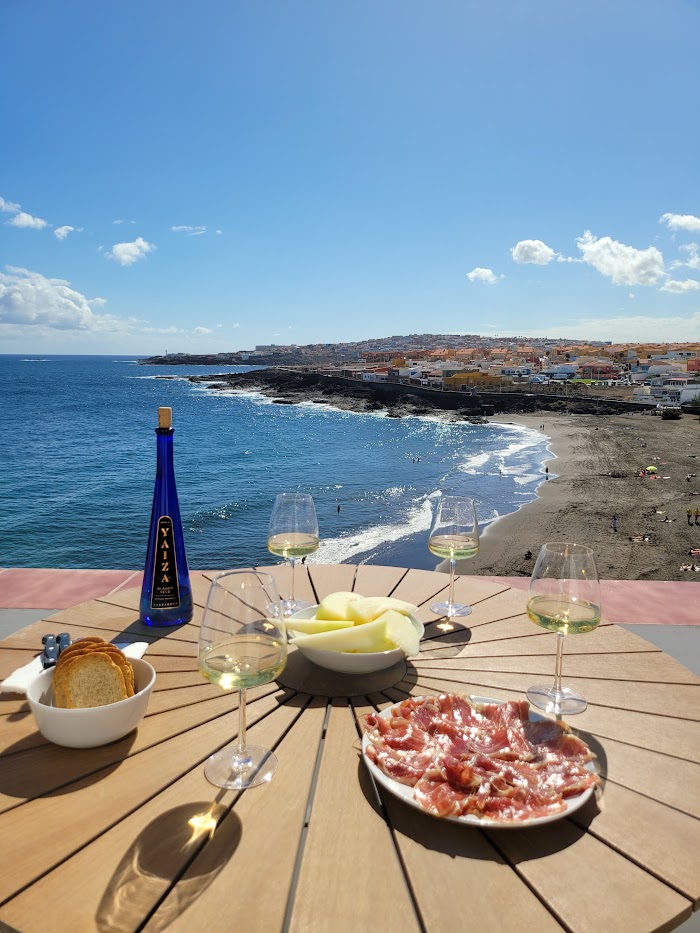 A dining table made of wood is set with glasses of white wine, fresh fruits, and a plate of sliced cured meat. In the background, a coastal view showcases clear blue water and a sandy beach, with colorful buildings dotting the hillside under a sunny sky.