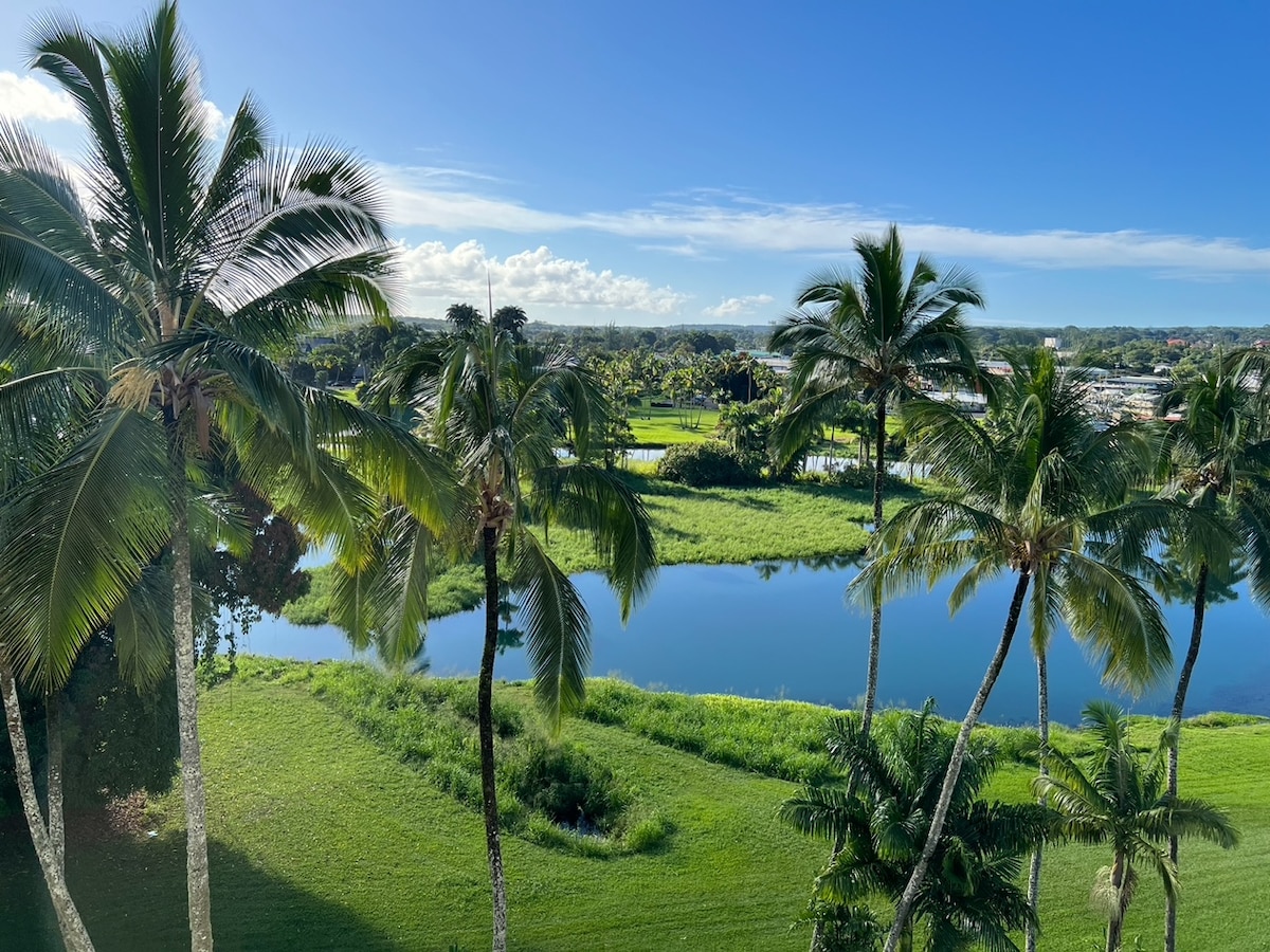 A vibrant view of palm trees frames a serene body of water, which reflects the blue sky. Lush green grass surrounds the scene, enhancing the natural beauty of the area. Distant trees and cloud formations add depth to the landscape, creating a peaceful atmosphere.