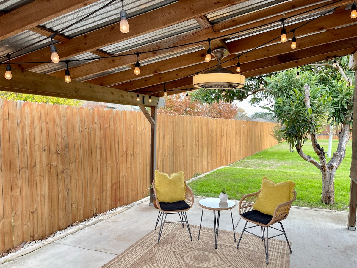 An outdoor seating area is presented with two chairs and a small side table. String lights hang overhead, providing soft illumination. A grassy area and a wooden fence can be seen in the background, contributing to a quiet and private setting.