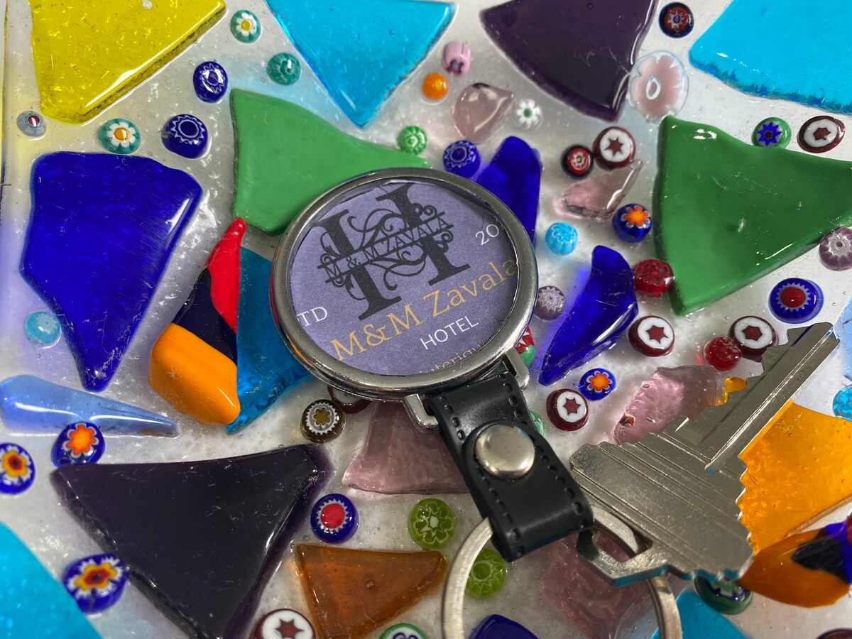 A hotel keychain rests on a vibrant mosaic surface made of multicolored glass pieces in various shapes and sizes. The keychain features a circular design with the text 'M&M Zaval Hotel' and the number '20', suggesting a unique and welcoming experience.