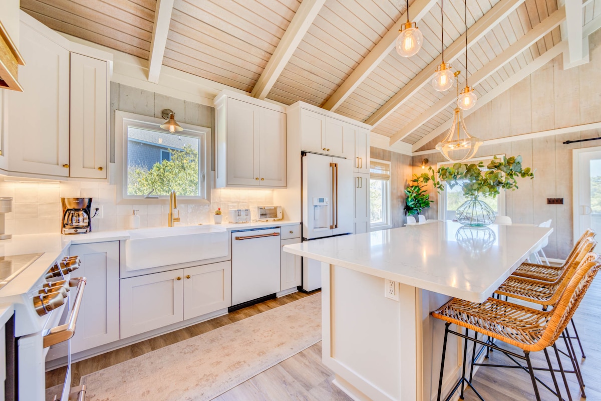 A modern kitchen is presented with spacious countertops and ample cabinetry. Stainless steel appliances are complemented by elegant light fixtures. A large island with bar seating for four invites social gatherings, while large windows allow natural light to illuminate the space.
