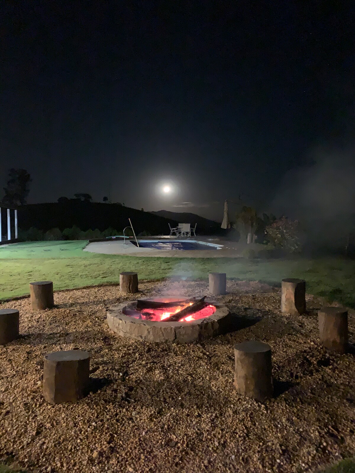 A cozy fire pit surrounded by wooden seating is illuminated by the light of the moon. Soft wisps of smoke rise gently into the night sky. In the background, a pool and lounge area are visible amidst the lush greenery.