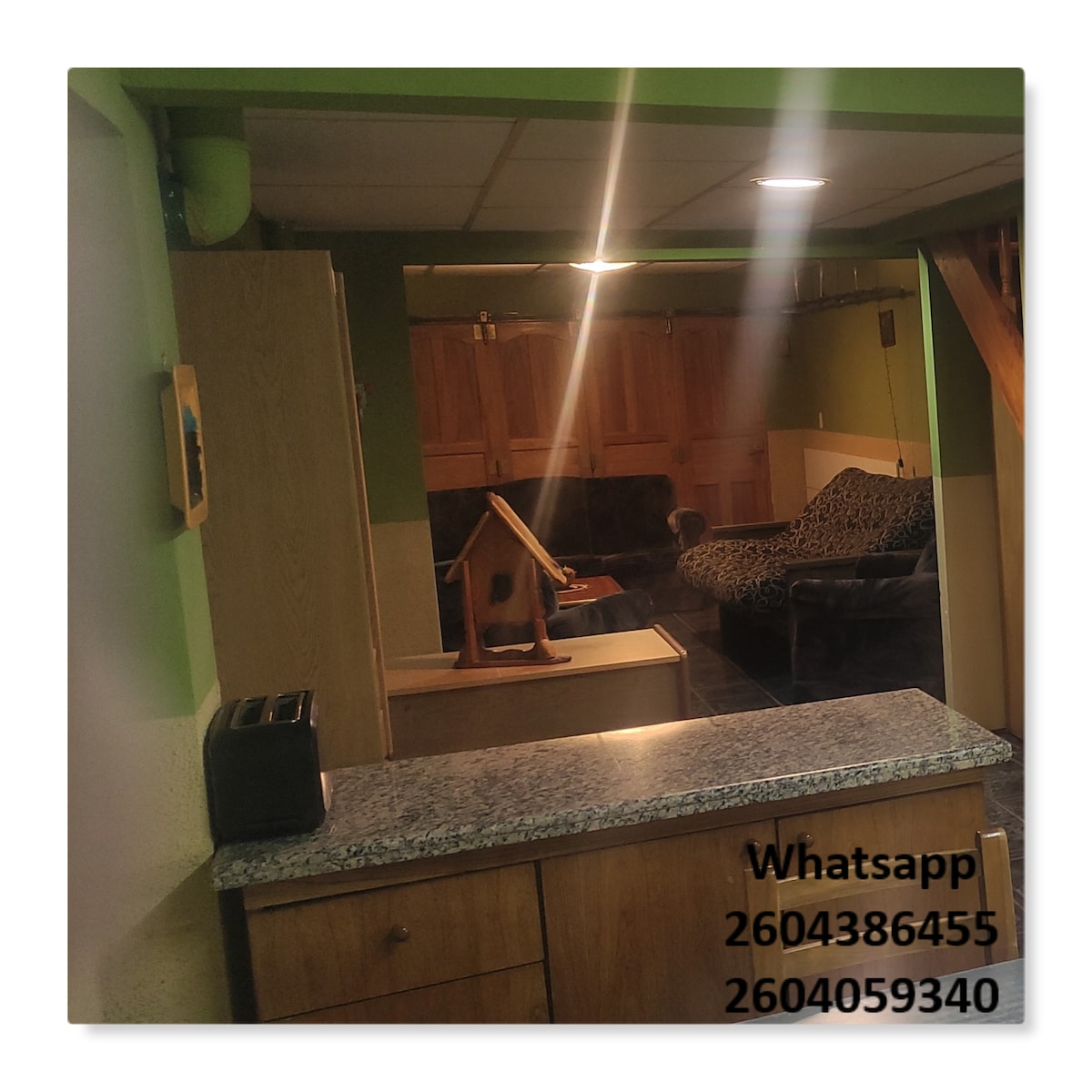 A view into the kitchen area reveals a countertop with a toaster and wooden cabinetry. Beyond, the living area is visible, featuring a spacious couch and a coffee table. The space is illuminated with warm lighting, highlighting the cozy ambiance.