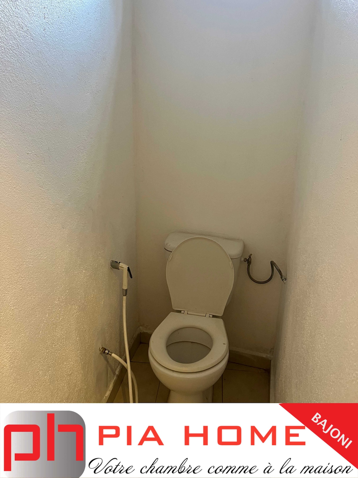 A modest bathroom space is presented, featuring a white toilet against a light-colored wall. The floor is tiled, and a plumbing fixture is visible next to the toilet, contributing to the functional design of the space.