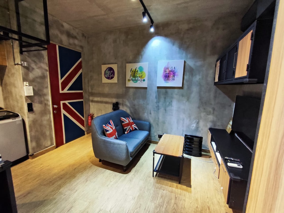 A cozy living area is highlighted by a gray sofa adorned with Union Jack-patterned cushions. Artistic wall prints in vibrant colors are displayed above a light wooden coffee table, while an entertainment unit and a kitchenette are visible in the background.