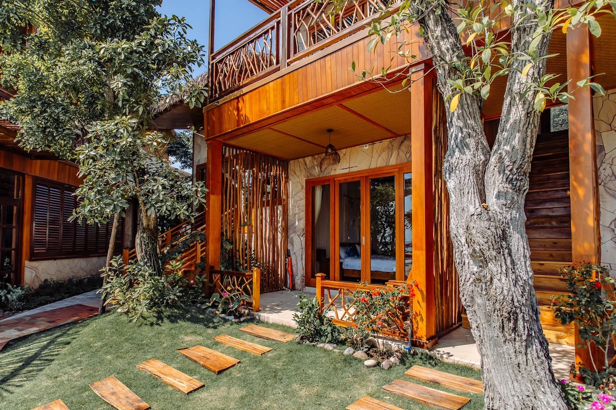 A rustic exterior of a wooden structure is framed by lush greenery. Pathways made of wooden planks lead through a manicured lawn toward a welcoming entrance, which is adorned with natural elements and large glass windows, creating a harmonious blend with the surrounding environment.
