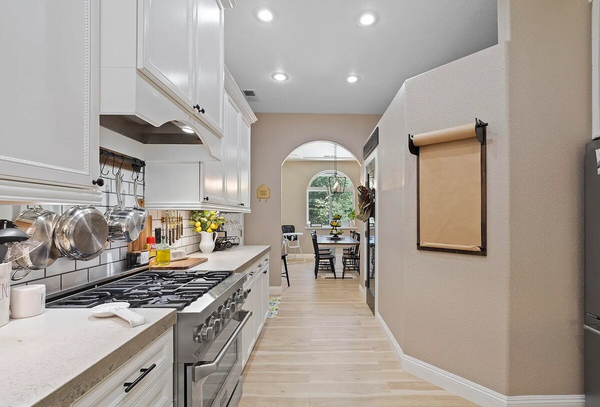 A modern kitchen features polished concrete countertops and a spacious layout. Stainless steel appliances are complemented by a row of hanging pots and pans. An arched window allows natural light to brighten the area, connecting to a cozy dining space with a wooden table and chairs.