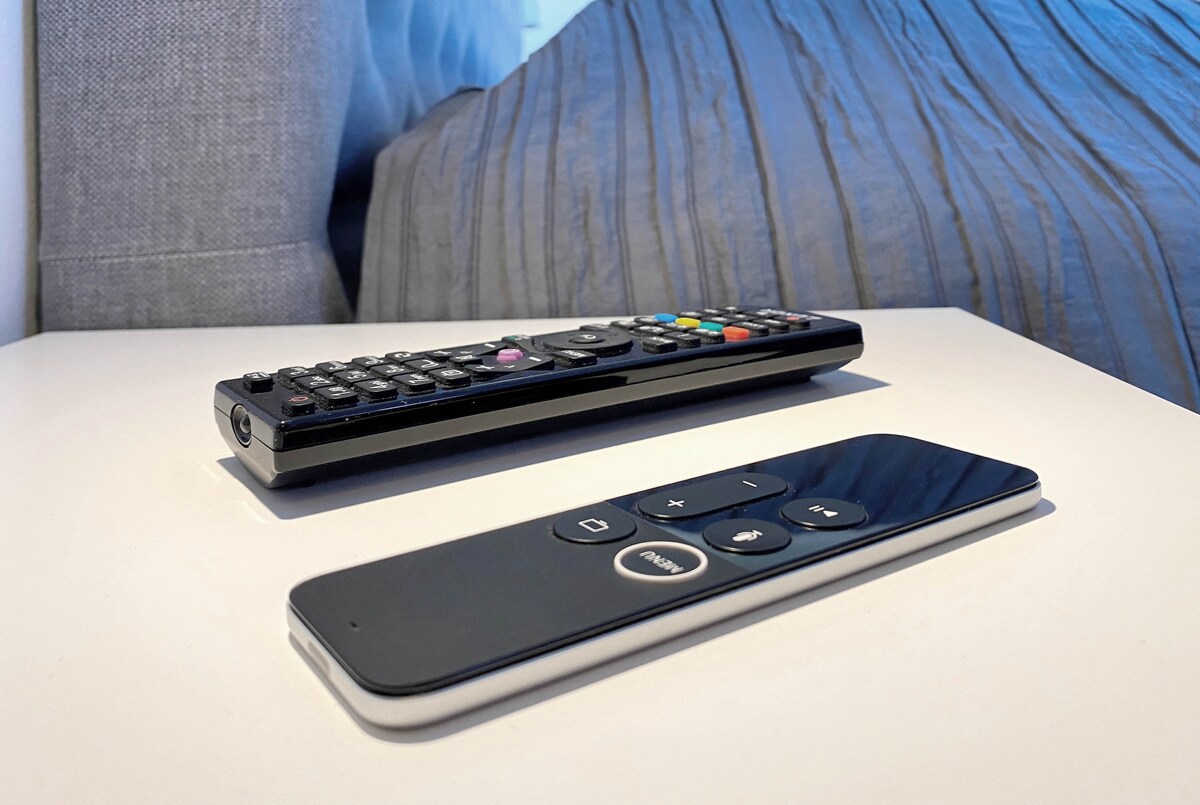 A bedside table is seen, featuring two remote controls positioned side by side. The background shows a neatly made bed with a textured gray bedspread, creating a sense of convenience and comfort.