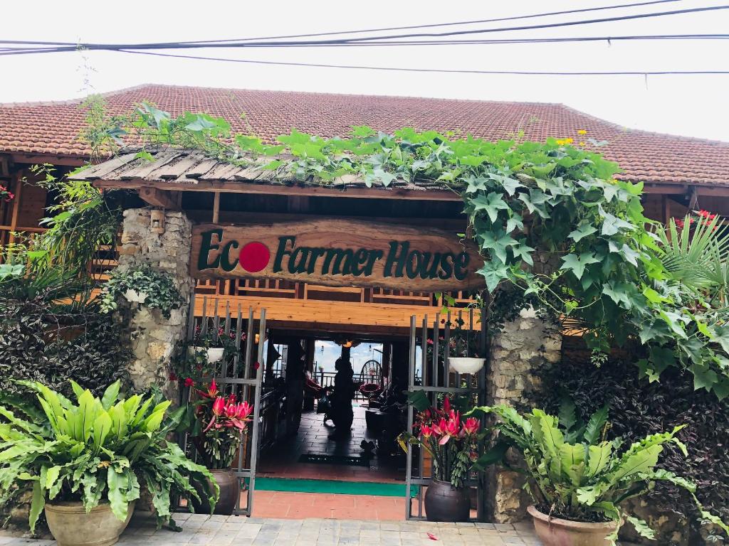 The entrance to Eco Farmer House features a welcoming sign surrounded by lush greenery and vibrant flowers. A stone pathway leads through decorative plants towards an open space within, where wooden beams and traditional architecture are complemented by natural elements.