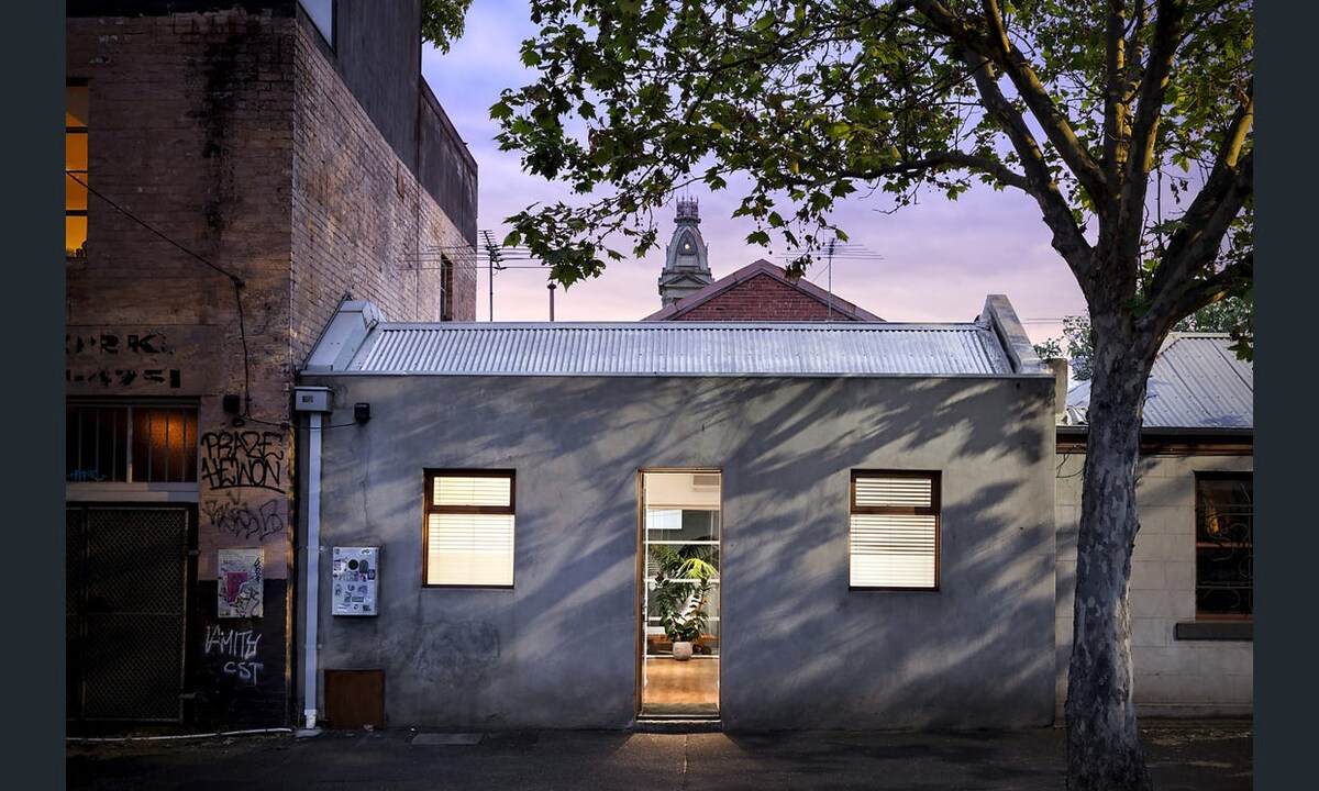 The exterior of a double fronted cottage is depicted, framed by a large tree. Two windows are visible on either side of the entrance, which is adorned with soft light. The building's textured wall contrasts with the setting dusk sky and iconic clock tower in the background.