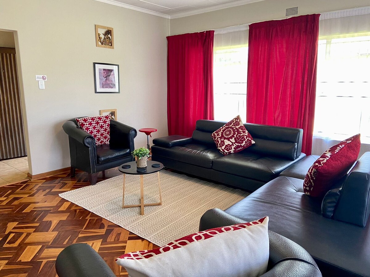 A comfortable living area features dark leather furnishings, including a sectional couch and armchair, arranged around a small, round table. Natural light filters through sheer curtains, enhancing the warm tones of the hardwood flooring and the rich red drapes.