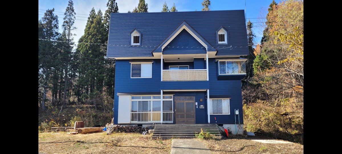 The exterior of a two-story blue mountain house is framed by tall trees. A spacious front porch with steps leads to the entrance, which is surrounded by large windows. The surrounding landscape features a grassy area, enhancing the home's connection to nature.
