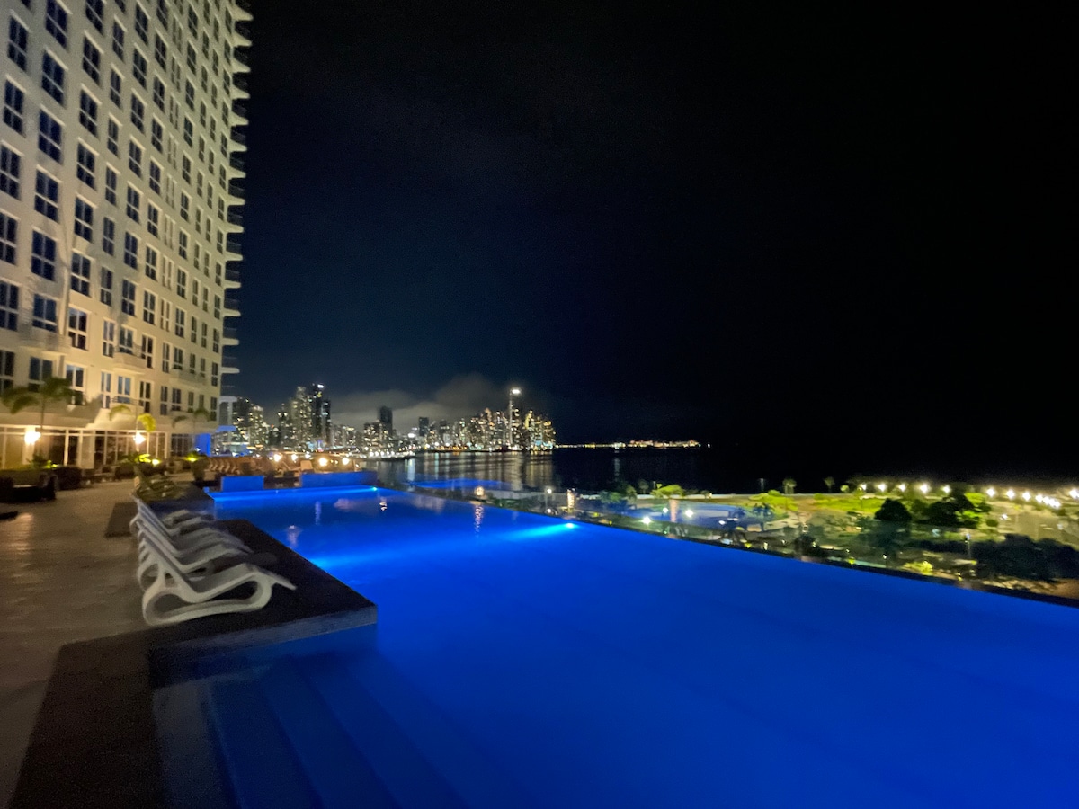 An infinity pool glows gently under the night sky, with sleek lounge chairs positioned at its edge. The skyline of the city is visible in the distance, illuminated by myriad lights reflecting on the water's surface.
