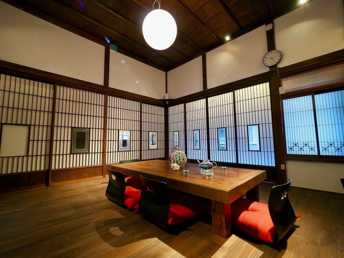 A spacious dining area features a large wooden table surrounded by four low chairs with red cushions. Traditional shoji screens allow natural light to filter through, highlighting the wooden beams and flooring. A round ceiling light provides soft illumination, complementing the room's serene atmosphere.