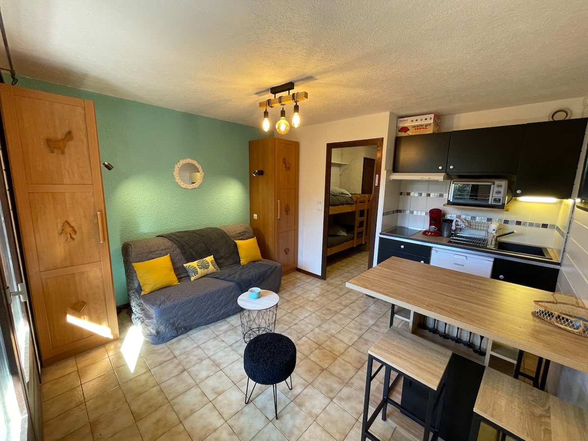 The open living area features a comfortable sofa with colorful cushions, complemented by a small dining table and black chairs. A kitchenette equipped with modern appliances is visible. Bright light enters through a window, illuminating the neutral-toned tile floor.