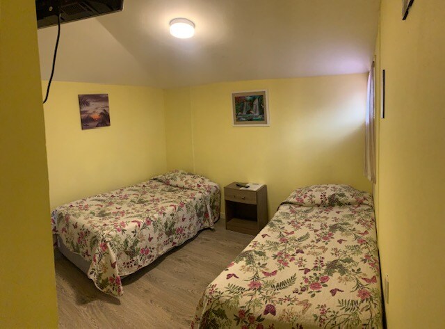 A bright room with yellow walls features two spacious beds, each covered with floral-patterned bedding. A nightstand is positioned between the beds, and natural light enters through a nearby window. Wall art adds a touch of color to the décor.