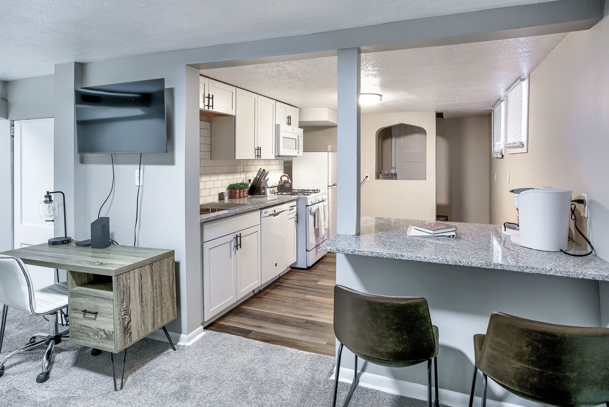 The image showcases a private studio featuring a modern kitchen with white cabinetry and stainless steel appliances. A workspace with a desk and chair is visible, along with a cozy seating area set against a granite countertop, creating a functional living space.