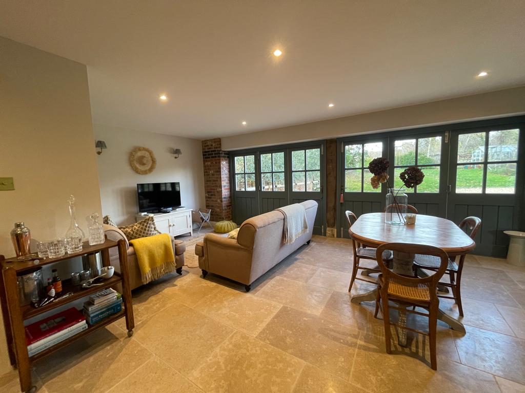 An open-plan living area features a comfortable sofa and a wooden dining table set for four. Panoramic windows provide views of the garden, allowing natural light to brighten the space. A drinks trolley with glassware and a cozy blanket adds to the inviting atmosphere.