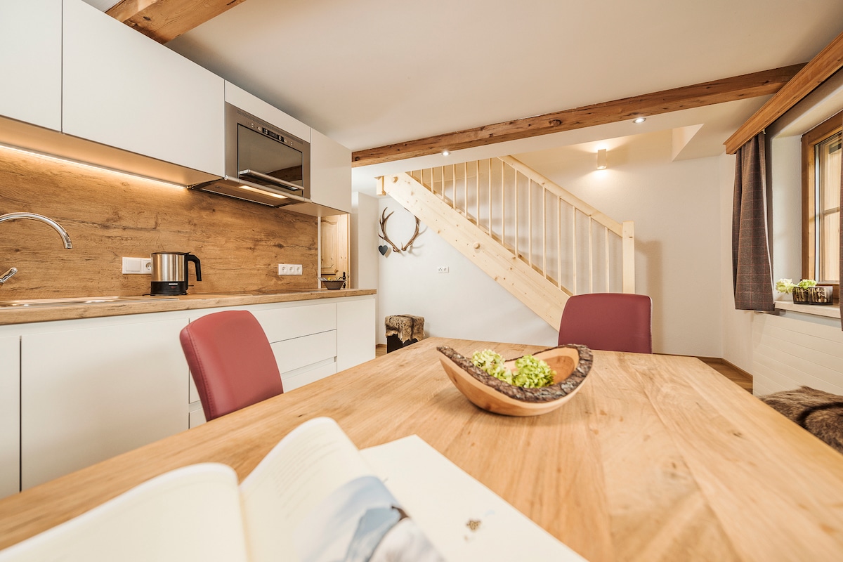 A well-equipped kitchen area features modern appliances and sleek cabinetry, complemented by a warm wooden dining table with red upholstered chairs. A staircase leads to the upper level, while decorative antlers add a rustic touch to the space.