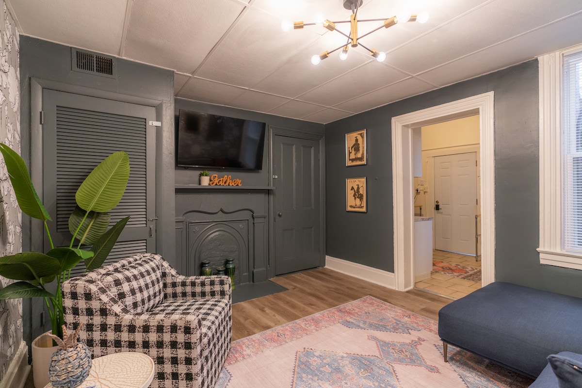 A cozy living area is highlighted by a checkered armchair and a spacious blue sofa. A light fixture with multiple bulbs hangs from the ceiling. A television is mounted on the wall, and decorative artwork is framed and displayed nearby.