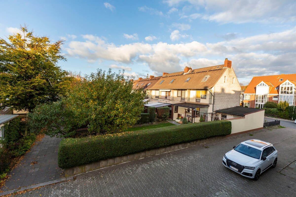 A peaceful residential neighborhood is depicted, featuring a line of multi-family houses set against a backdrop of scattered clouds. A neatly trimmed hedge borders the driveway, and a parked car is visible on the cobblestone street, while trees add greenery to the scene.