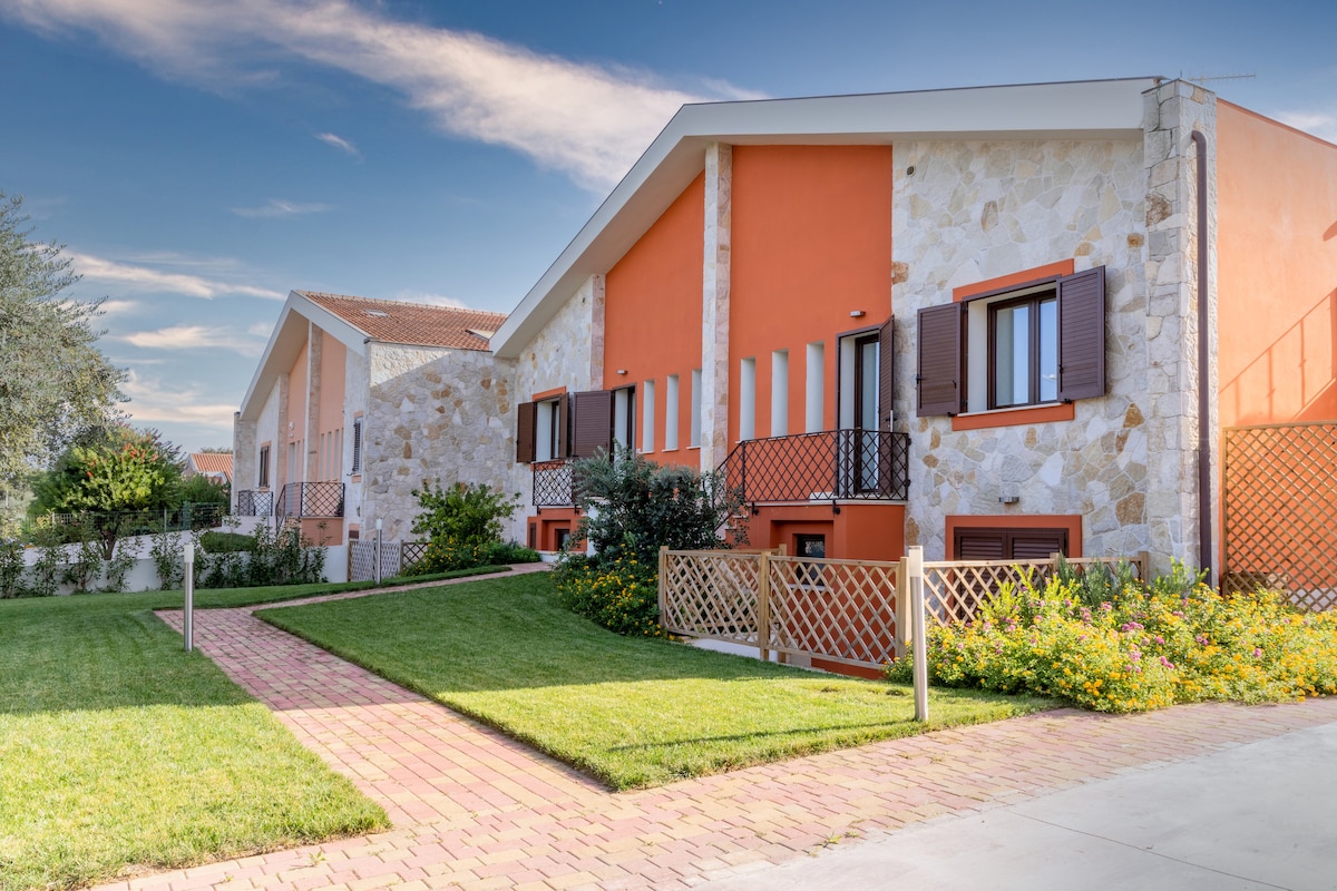A modern building complex features a combination of stone and stucco facades, highlighted by vibrant orange accents. Well-maintained gardens and pathways lead to the entrance, surrounded by lush greenery and colorful flower beds, providing a welcoming outdoor space.