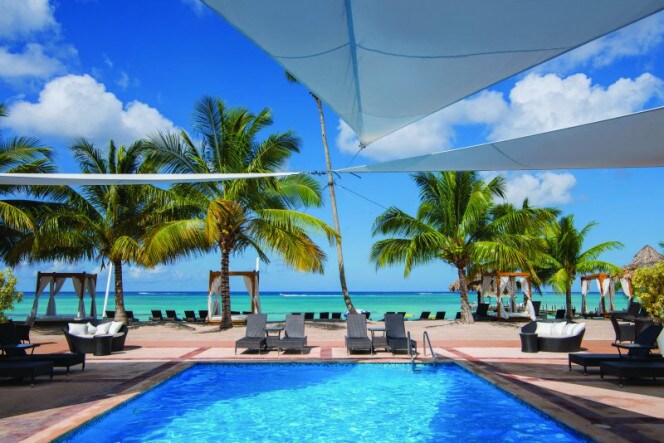 A pool area is framed by tall palm trees, with sun loungers arranged nearby. White canopies provide shade over the blue water of the pool, reflecting the clear sky above. In the background, the beach meets the turquoise sea.
