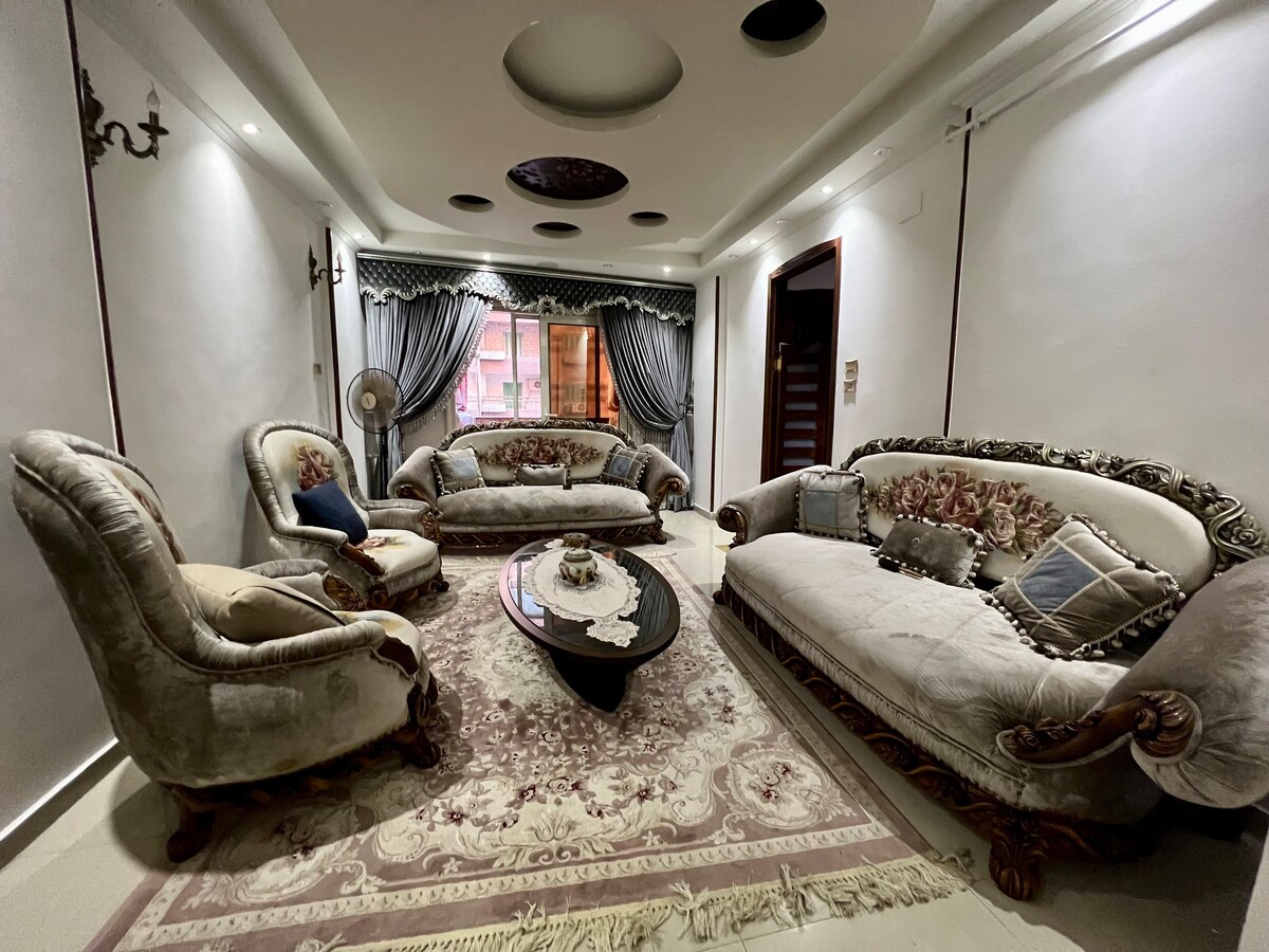 A spacious living room is presented with elegant furniture, including a plush sofa set and a decorative armchair. A circular coffee table sits on a patterned rug, while large windows draped in curtains allow natural light to fill the space.