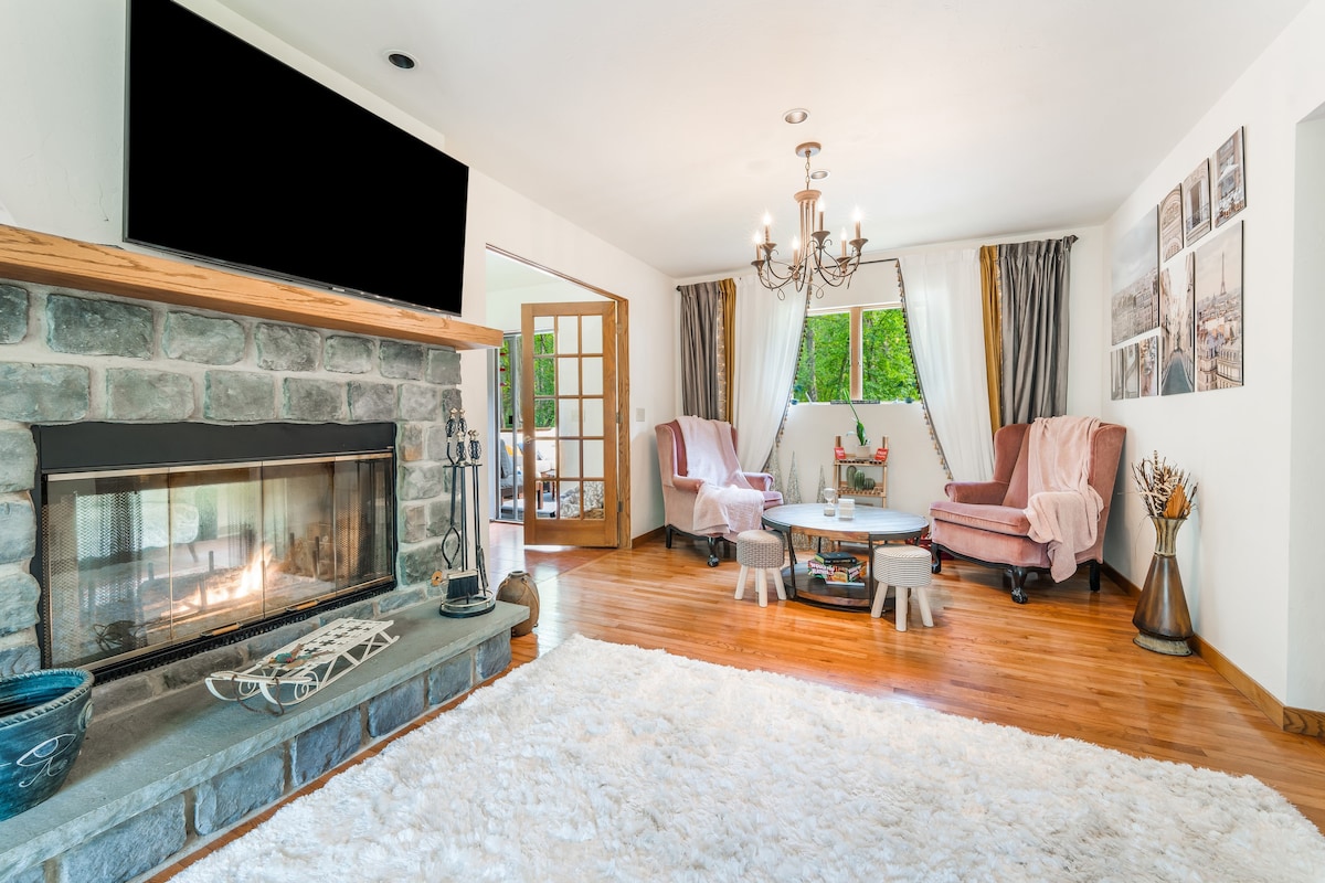A spacious living area is highlighted by a stone fireplace and warm wooden floors. Two cozy armchairs are arranged around a central table, creating a welcoming atmosphere. Soft white carpeting extends across the floor, while large windows allow natural light and views of the outdoors.