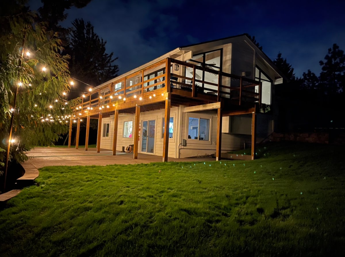 A modern farmhouse's exterior is illuminated by soft string lights, casting a warm glow on the spacious wooden deck. The large windows reflect the evening sky, while the lush green lawn provides a serene atmosphere, creating an inviting space for outdoor relaxation.
