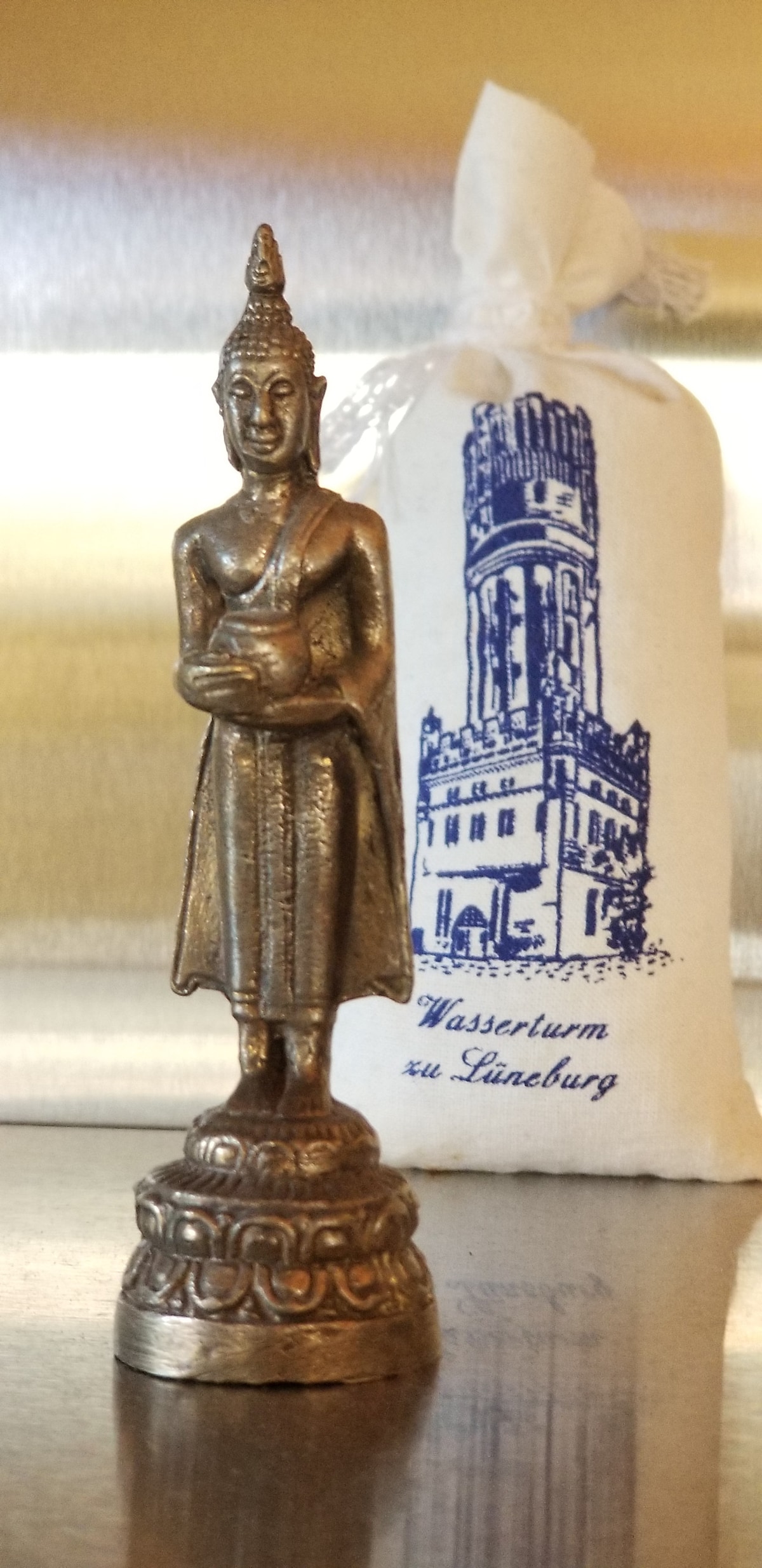 A small metallic statue of a standing figure is displayed on a reflective surface, showcasing intricate detailing on clothing and a serene expression. In the background, a white pouch features a blue illustration of a historic tower, providing a contrast to the statue.