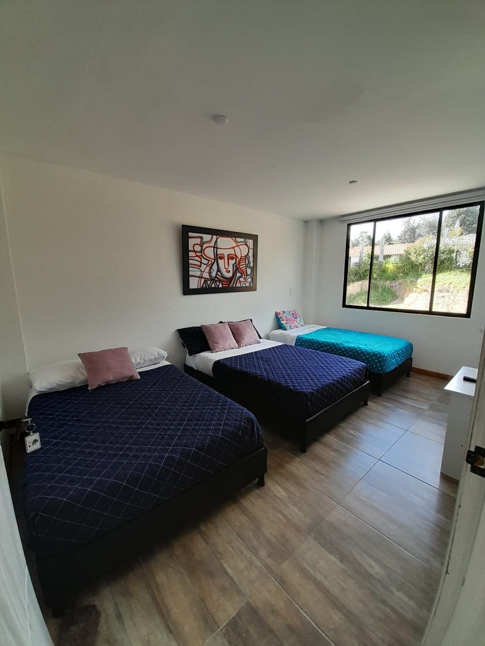 A thoughtfully arranged guest room is presented with three spacious beds, each adorned with patterned bedding. A large window allows natural light to brighten the space, revealing a view of greenery outside. A piece of wall art adds a touch of color to the decor.