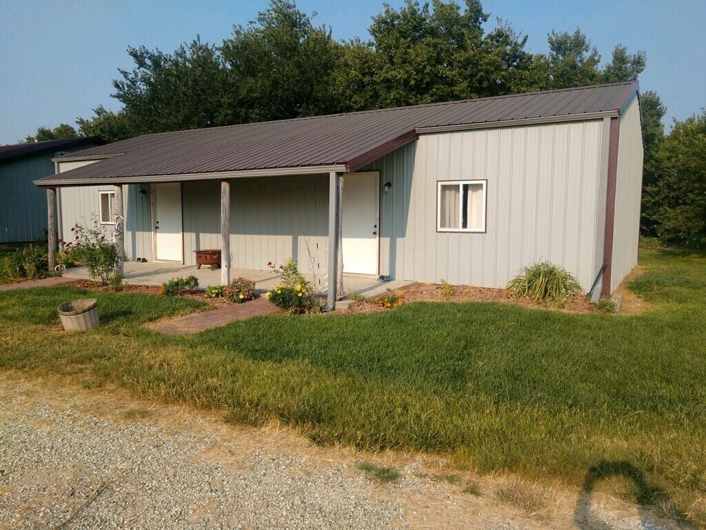 A simple structure features a metal roof with light-colored siding, set in a grassy area. A covered porch is visible with a small bench and flowering plants along the pathway. Trees and greenery surround the building, contributing to a serene atmosphere.