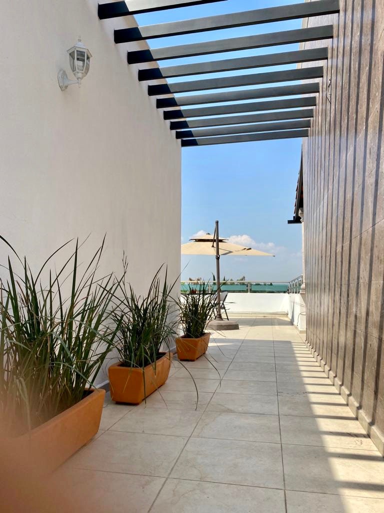 A tiled pathway is flanked by potted plants, leading towards a serene view of the ocean. An umbrella stands beside a seating area in the distance, while natural light filters through the overhead structure, casting shadows on the ground.