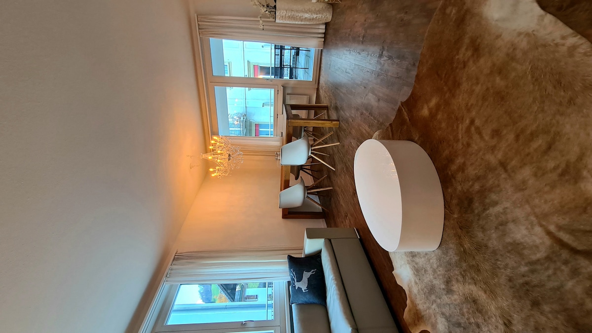 A bright living area features large windows allowing natural light to fill the space. A cozy, neutral-toned sofa is paired with a round coffee table. Two stylish chairs are positioned at a dining table, complemented by a decorative light fixture above.