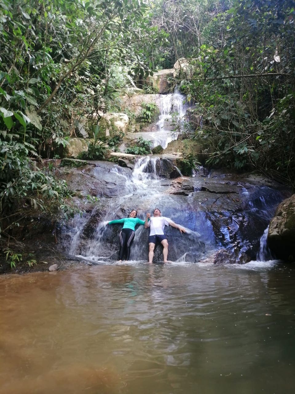 A serene natural setting features two individuals relaxing beside a small waterfall cascading over rocks into a calm pool. Lush greenery surrounds the area, enhancing the tranquil environment, with sunlight filtering through the trees above.