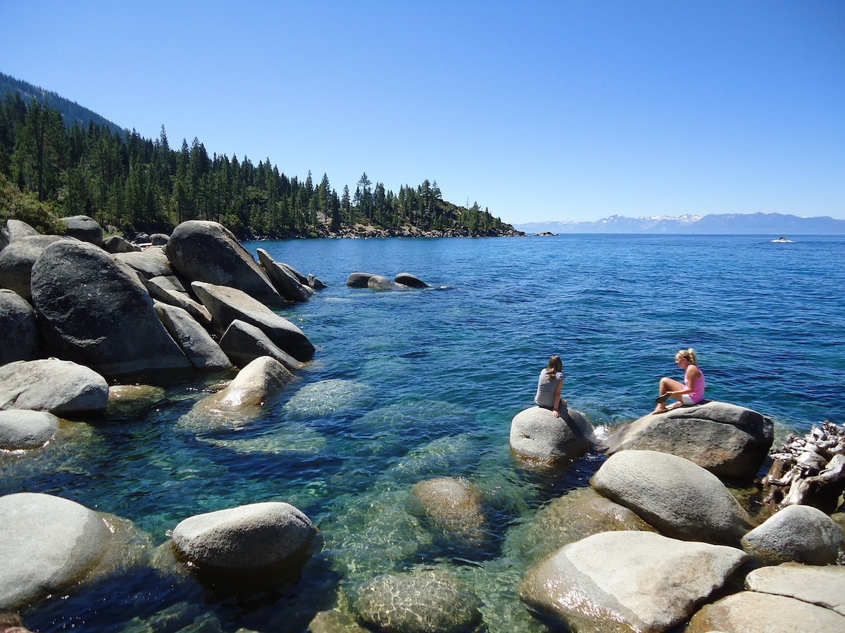 Clear blue water reflects the sky near the shoreline of a lake, with large smooth rocks scattered along the edge. Two individuals are seated on the rocks, enjoying the view of the surrounding pine trees and distant mountains.
