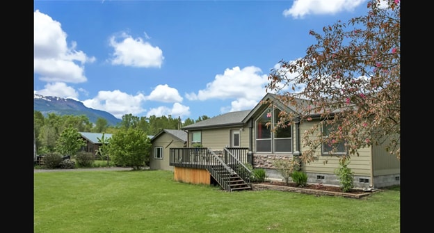 The cottage exterior is framed by vast green lawn, with a wooden deck leading to the entrance. Large windows allow light into the living areas, while mountains are visible in the background. The landscape includes trees, adding to the serene atmosphere.