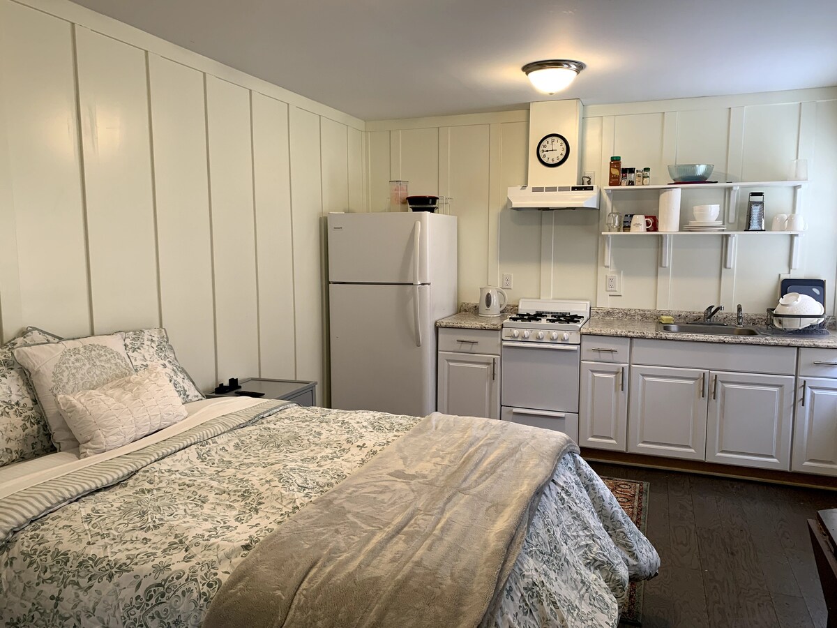 A cozy sleeping area features a full-sized bed with a patterned comforter, complemented by bedside tables. A kitchenette, equipped with a refrigerator, stove, and open shelving, is visible in the background. Natural light brightens the space, highlighting wooden flooring and white wall paneling.