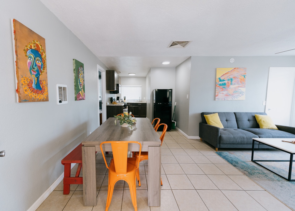 A spacious dining area showcases a wooden table surrounded by colorful chairs. Bright artwork adorns the walls, adding a touch of vibrancy. A well-equipped kitchen is visible in the background, with modern appliances and a dark color scheme contrasting the light tile floor.