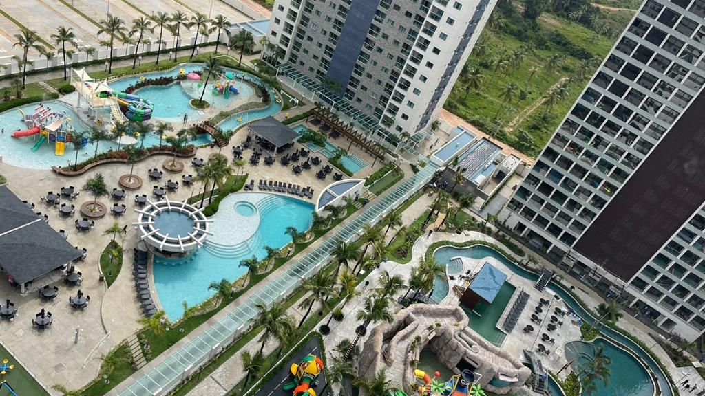 An expansive outdoor recreational area is depicted, featuring multiple swimming pools surrounded by lounge chairs. Brightly colored water play structures for children are visible, alongside lush greenery and palm trees. The layout includes shaded seating areas, providing ample space for family enjoyment.