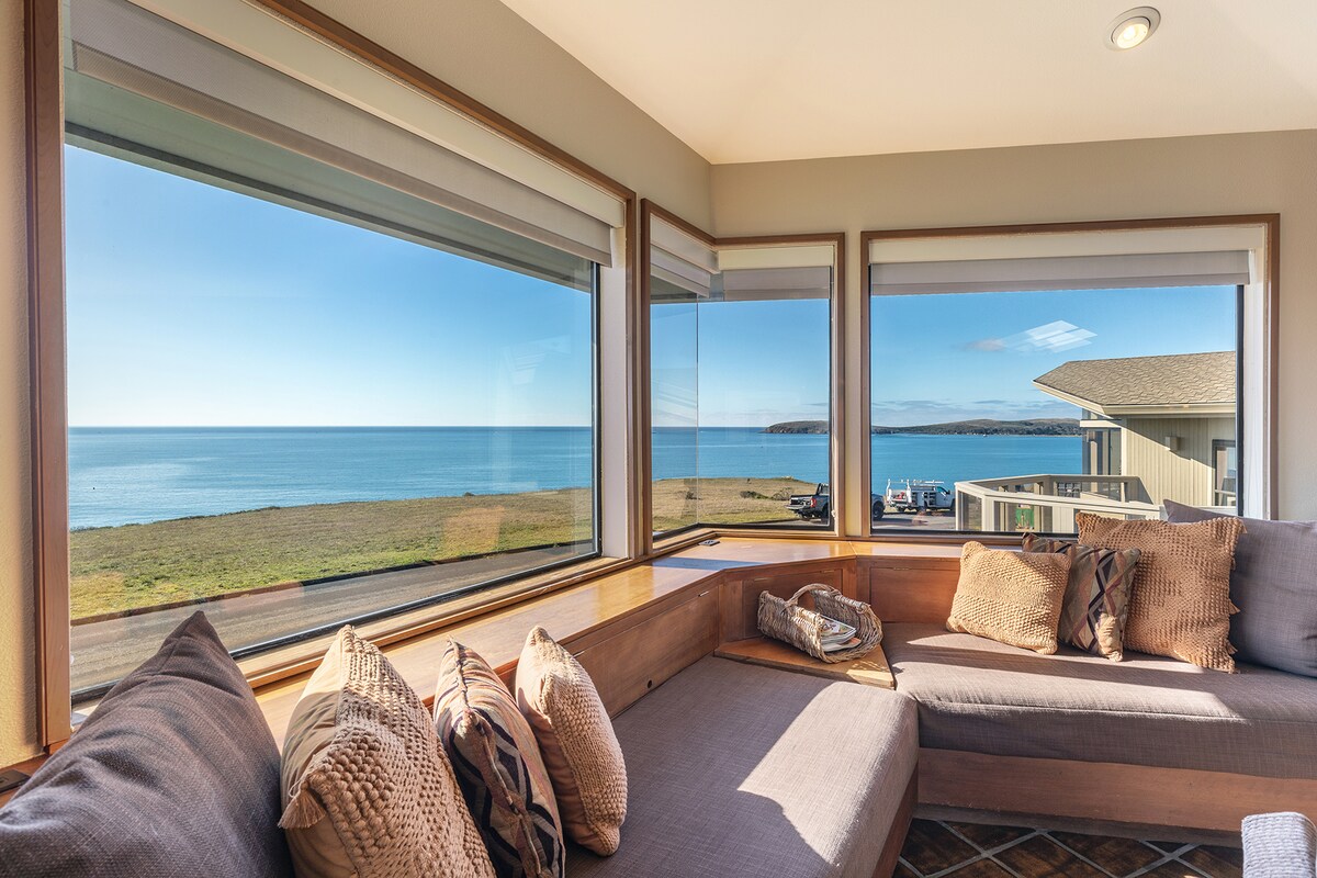 A comfortable seating area features a sectional sofa adorned with textured pillows. Large windows frame unobstructed views of the ocean and coastline, allowing natural light to fill the space. The serene blue waters and clear skies create a peaceful ambiance.