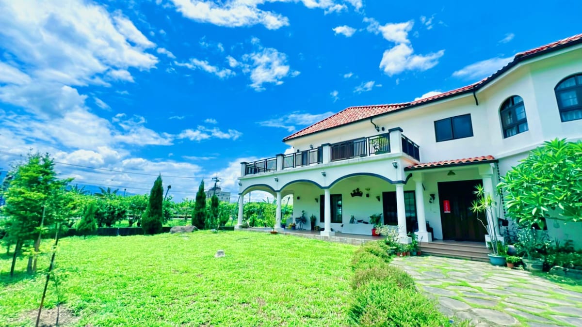 A two-story house is set against a bright blue sky, featuring a terracotta-tiled roof and white exterior walls. Lush greenery surrounds the property, including small trees and a well-maintained lawn, while a stone pathway leads to the entrance, inviting a sense of tranquility.